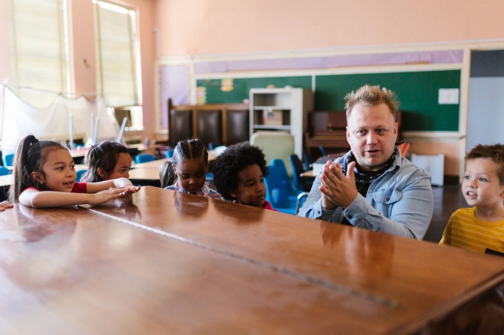 A teacher interacts with diverse children around a desk in a classroom setting. — Challenges to Overcome